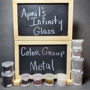 Puede incluir: Una colección de pequeños frascos de vidrio llenos de cuentas de vidrio de diferentes colores. Los frascos están etiquetados con nombres como "Smoke", "Black", "Tarnished Gold", "Medallion Gold", "Cloudy", "Alloy Sterling", "Gun Metal" y "Sterling Silver". Los frascos están dispuestos en una fila sobre una superficie negra. Los frascos están etiquetados con las palabras "April's Infinity Glass" y "Color Group Metal".