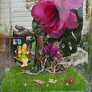 May include: A miniature fairy garden terrarium featuring a pink flower, a green fairy sitting on a swing, and green grass.