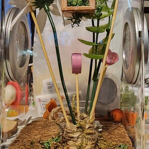 May include: A clear glass terrarium with a brown base and a lid. Inside the terrarium, there are several artificial plants, including a white flower with pink accents, green leaves, and a small wooden house with a green moss roof. There are also pink and purple decorative accents on wooden sticks.