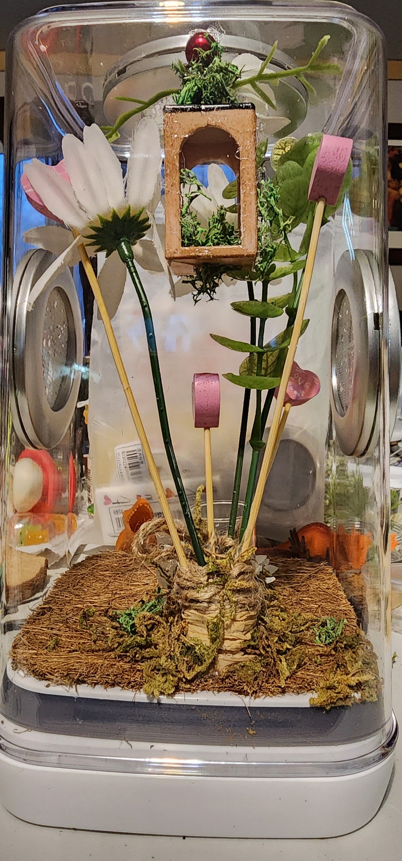 May include: A clear glass terrarium with a brown base and a lid. Inside the terrarium, there are several artificial plants, including a white flower with pink accents, green leaves, and a small wooden house with a green moss roof. There are also pink and purple decorative accents on wooden sticks.
