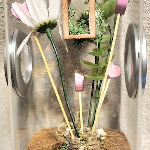 May include: A clear glass jar terrarium with a brown base and a white lid. Inside the jar are green plants, a small wooden house, and pink hearts on sticks.