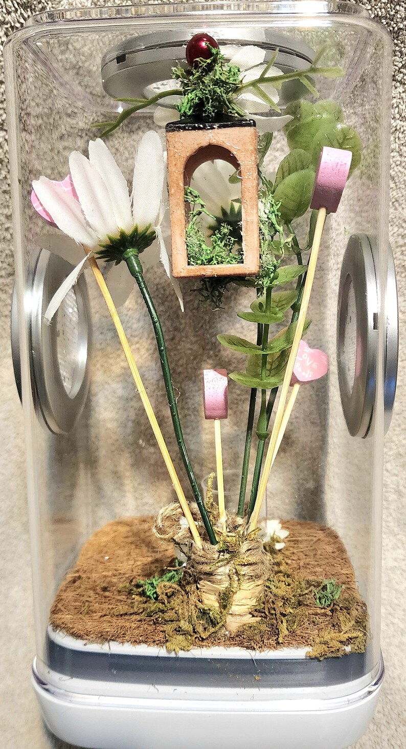 May include: A clear glass jar terrarium with a brown base and a white lid. Inside the jar are green plants, a small wooden house, and pink hearts on sticks.
