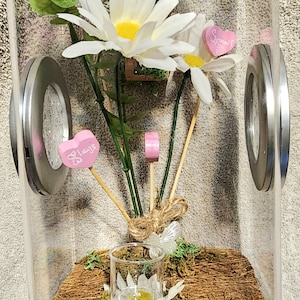 May include: A clear glass terrarium with a white base, containing a small potted plant with white flowers, a small glass jar with a white flower, and two pink wooden hearts with the words "Space" and "Hugs" written on them.