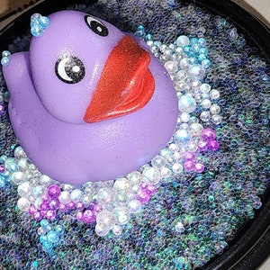 May include: A purple rubber ducky with a red beak and black eyes sits in a bed of iridescent blue, white, and pink glitter.