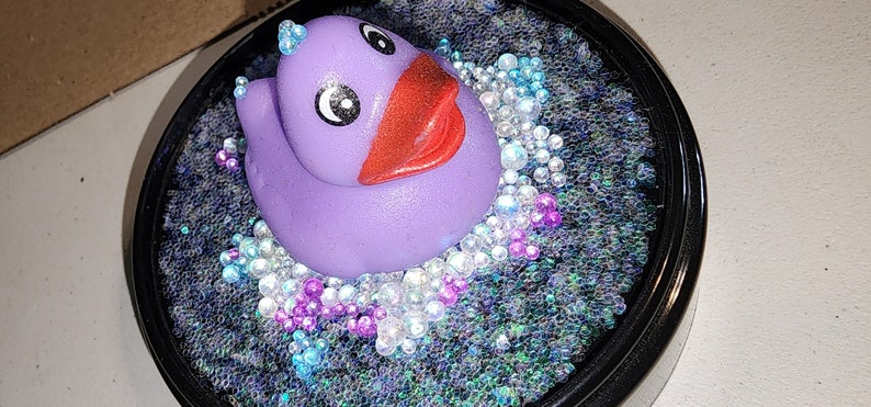 May include: A purple rubber ducky with a red beak and black eyes sits in a bed of iridescent blue, white, and pink glitter.