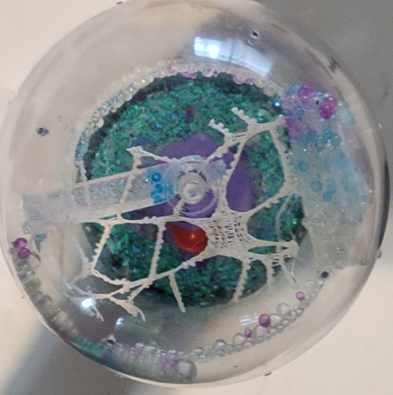 May include: A clear plastic sphere filled with iridescent green glitter, white lace, and purple and blue beads. A small red object is visible in the center of the sphere.