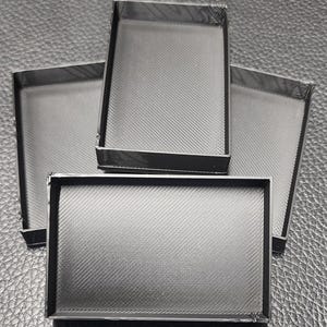 May include: Four rectangular, open-top trays with a textured, diagonal pattern. The trays are dark gray with black trim, arranged on a dark surface. The trays appear to be made of a rigid material.