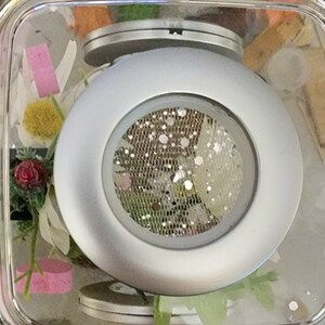 May include: A silver-colored round object with a mesh screen in the center. The object is in a clear container with other decorative items.