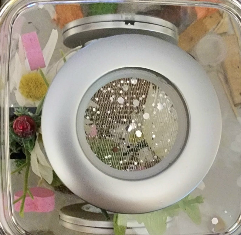 May include: A silver-colored round object with a mesh screen in the center. The object is in a clear container with other decorative items.