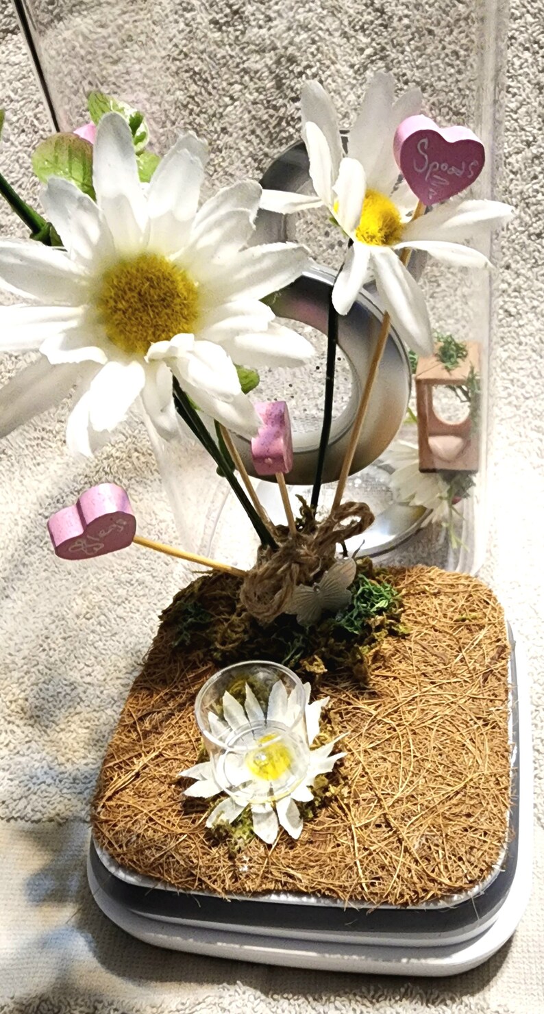 May include: A clear glass jar with a white lid, filled with a floral arrangement of white daisies and pink hearts. The jar is sitting on a brown coir mat.