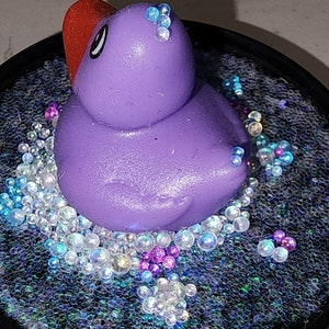 May include: A purple rubber ducky with a red bill sits in a clear resin circle filled with iridescent glitter and small, clear beads.