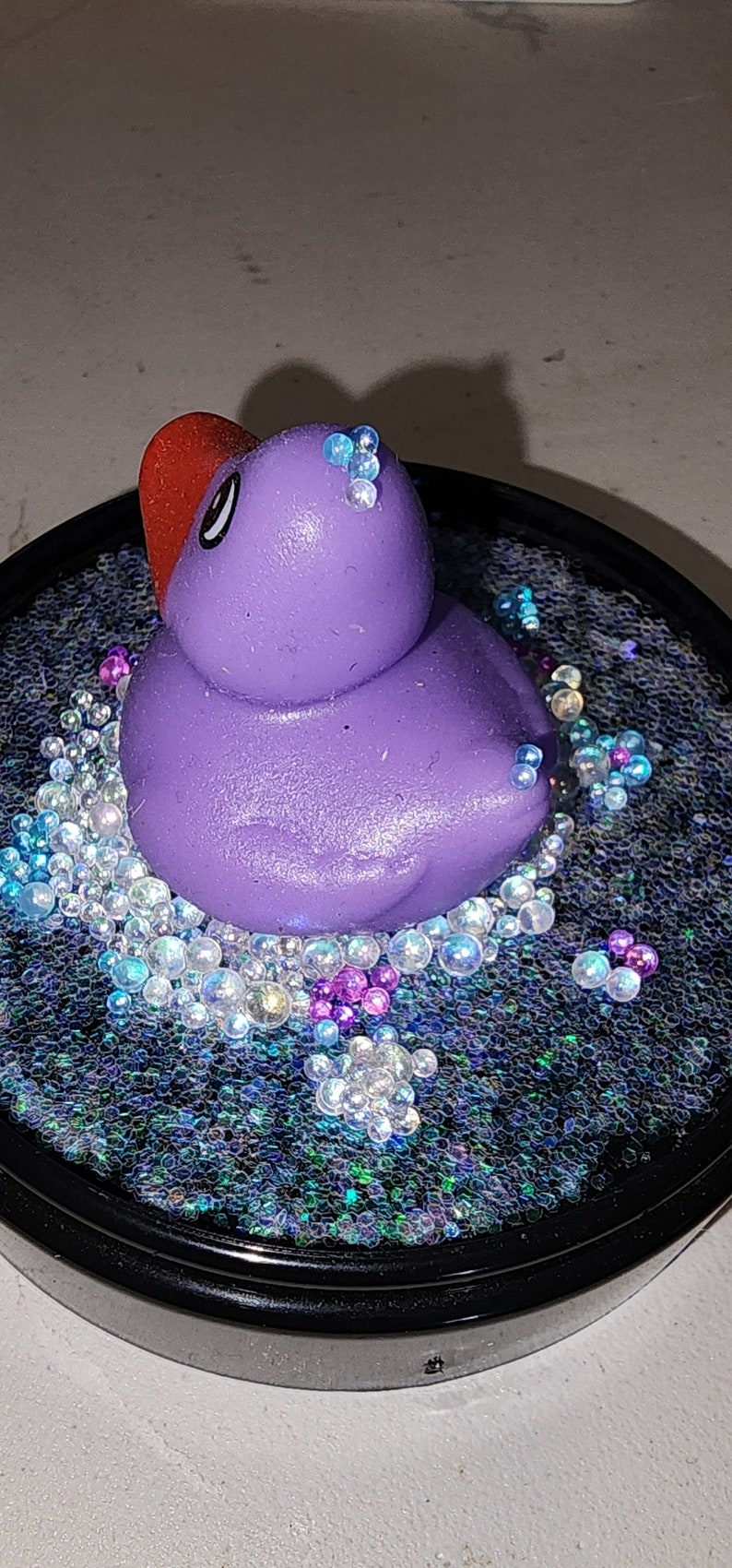May include: A purple rubber ducky with a red bill sits in a clear resin circle filled with iridescent glitter and small, clear beads.