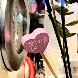 May include: A pink heart-shaped candy with the word "bless" written on it. The candy is in a glass jar with other decorative items.