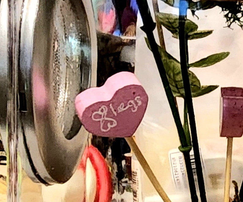 May include: A pink heart-shaped candy with the word "bless" written on it. The candy is in a glass jar with other decorative items.