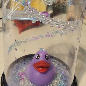 May include: A purple rubber ducky sits in a glass dome filled with iridescent glitter and small, clear beads. The dome has a black base.