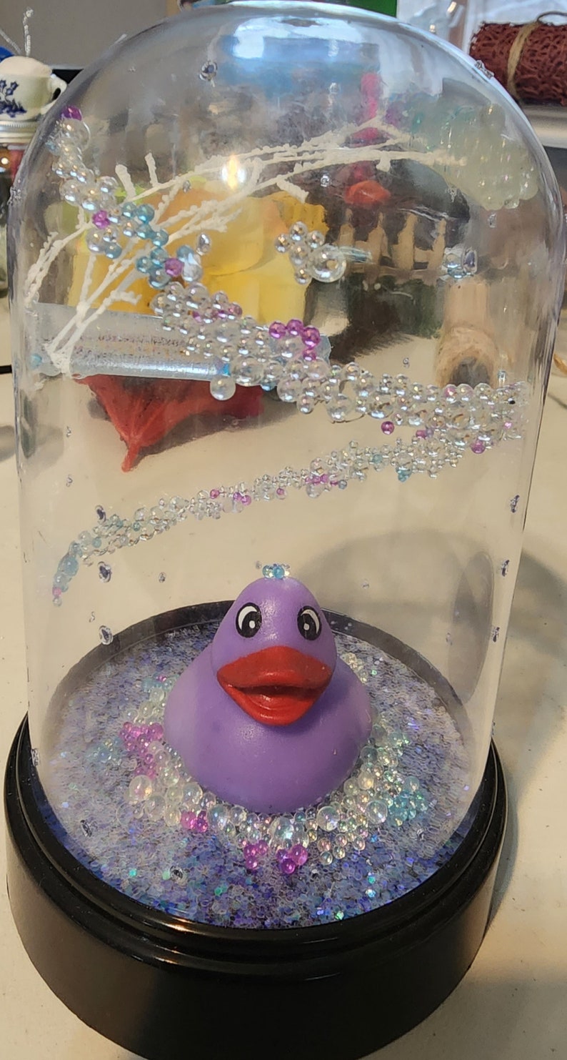 May include: A purple rubber ducky sits in a glass dome filled with iridescent glitter and small, clear beads. The dome has a black base.