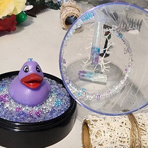 May include: A purple rubber ducky sits in a clear plastic container filled with iridescent blue and purple beads. The container has a black rim and a clear lid with a white snowflake design.