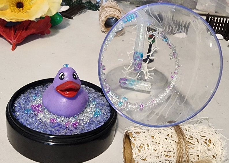 May include: A purple rubber ducky sits in a clear plastic container filled with iridescent blue and purple beads. The container has a black rim and a clear lid with a white snowflake design.