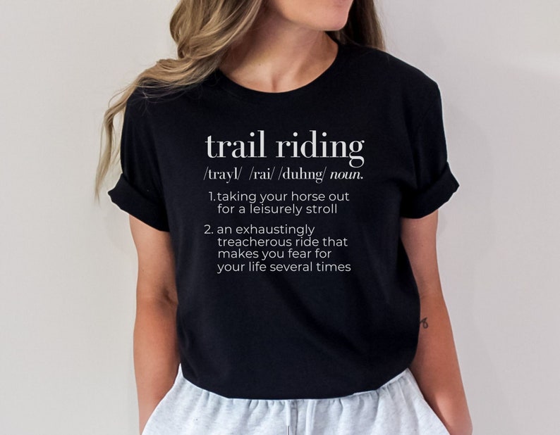 May include: Black t-shirt featuring a humorous definition of 'trail riding.'  The definition includes two points: a leisurely stroll and an exhausting, treacherous ride. The text is white and uses a dictionary-style format. The shirt is shown on a model, but the focus is on the shirt's design.