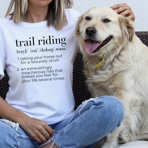 May include: White t-shirt with the text "trail riding" defined as "taking your horse out for a leisurely stroll" and "an exhaustingly treacherous ride that makes you fear for your life several times". A person wearing the shirt is petting a golden retriever.