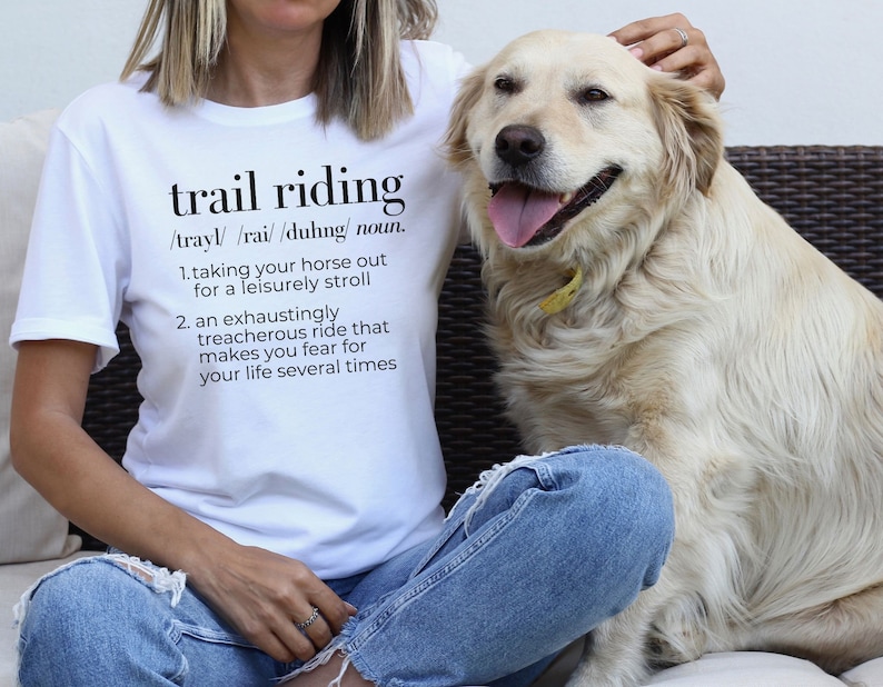 May include: White t-shirt with the text "trail riding" defined as "taking your horse out for a leisurely stroll" and "an exhaustingly treacherous ride that makes you fear for your life several times". A person wearing the shirt is petting a golden retriever.