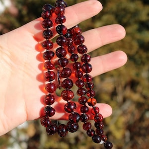 May include: A necklace of dark red amber beads. The beads are round and vary in size, strung together on a dark string. The necklace is held in a hand, with the beads cascading down. The amber has a glossy finish.