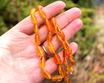 May include: A necklace of amber-colored beads. The beads are oval-shaped and strung together. The necklace is held in a hand, with a blurred green background.