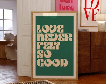 Puede incluir: Una gran pieza de arte mural rectangular enmarcada con un fondo verde oscuro y el texto beige "LOVE NEVER FELT SO GOOD" en una tipografía audaz de estilo retro. La impresión se muestra en un marco de madera clara sobre el suelo.