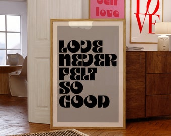 Puede incluir: Una pieza de arte mural rectangular enmarcada que presenta un fondo gris con el texto negro "LOVE NEVER FELT SO GOOD" en una fuente negrita y estilizada. El marco es de madera clara y la impresión se muestra en orientación vertical.