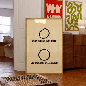 May include: A framed poster with a beige background and black text that reads "JUST MAKE IT EXIST FIRST" and "YOU CAN MAKE IT GOOD LATER". The poster has two black circles and a cloud graphic. Other framed art is visible in the background.