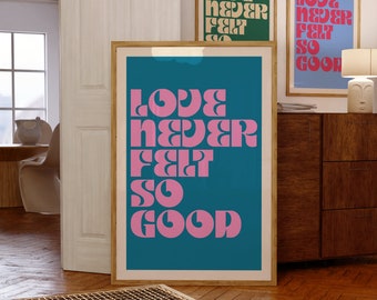 Puede incluir: Una lámina de arte mural vertical enmarcada con un fondo azul verdoso intenso y el texto "LOVE NEVER FELT SO GOOD" en una fuente serif rosa, audaz y estilizada. La lámina se presenta en un marco de madera clara, con otras dos láminas visibles detrás.