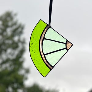 May include: A stained glass lime slice suncatcher with a bright green rind and light green segments. The glass is outlined with black and has a brown border. A black ribbon is attached for hanging.