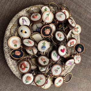 May include: A collection of handmade embroidered keyrings featuring various floral designs. The keyrings are made with wooden hoops and feature colourful embroidery on white fabric.