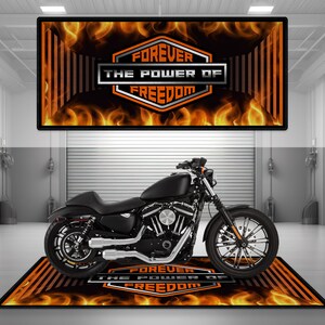 May include: Black motorcycle with chrome detailing, positioned on a mat featuring the text "FOREVER THE POWER OF FREEDOM" and a flame design. A matching sign is above. The setting is a garage.