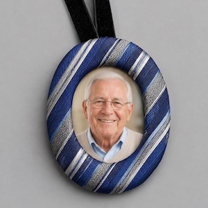 May include: An oval-shaped memorial ornament featuring a portrait of an elderly person with white hair and glasses. The frame is wrapped in a blue, silver, and white striped fabric, likely repurposed from a necktie, and includes a black ribbon hanger.