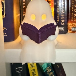 May include: A white, translucent ghost figurine with hollow, glowing yellow eyes. The ghost is holding an open, dark purple book in its hands. The figure has a smooth, rounded head and a draped, flowing body shape that sits upright on a flat surface.