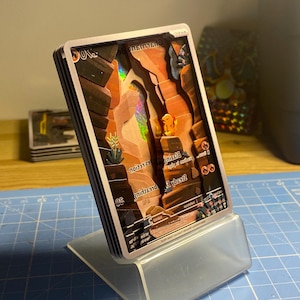 May include: A collection of trading cards depicting a desert canyon, presented in a clear acrylic stand. The cards have a holographic effect and are on a blue grid surface.