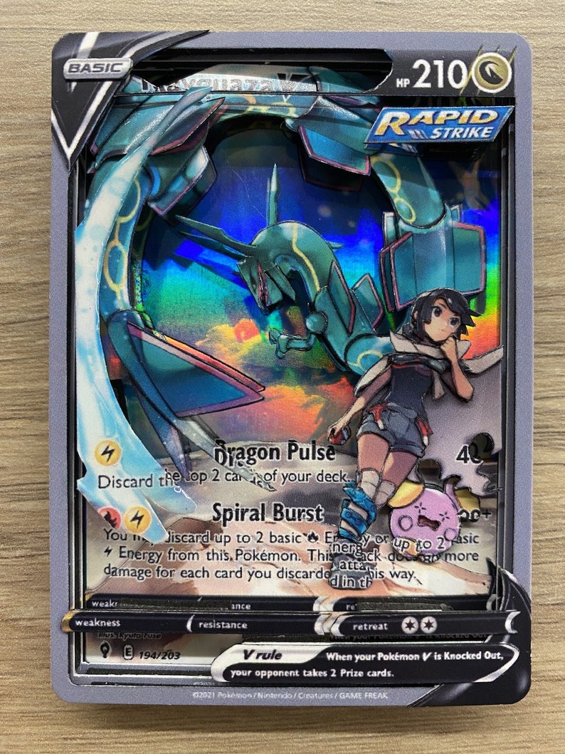 May include: A Pokemon trading card featuring a green and blue dragon Pokemon with a trainer. The card is a basic card with a rapid strike ability. The card has a dragon pulse attack that discards the top 2 cards of your deck and a spiral burst attack that discards up to 2 basic energy cards or up to 2 basic energy cards from this Pokemon. The card has a V rule that states when your Pokemon V is knocked out, your opponent takes 2 prize cards. The card is number 194/203.
