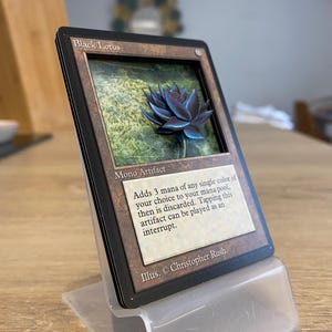 Peut inclure: Une carte "Black Lotus" encadrée, un Mono Artifact, avec une fleur de lotus bleue en 3D. La carte est présentée dans un présentoir en plastique transparent. Le texte sur la carte indique "Adds 3 mana..." et "Illus. © Christopher Rush."