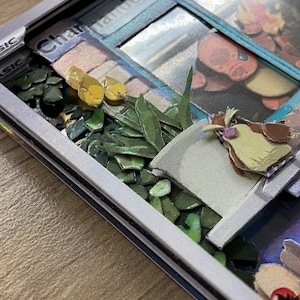 May include: A close-up of a Pokemon card with a white border and the word 'BASIC' in black text. The card features a 3D image of a Charmander Pokemon in a window with a green plant and a brown bird in the foreground. The card is on a wooden surface.