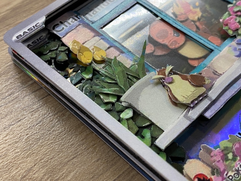 May include: A close-up of a Pokemon card with a white border and the word 'BASIC' in black text. The card features a 3D image of a Charmander Pokemon in a window with a green plant and a brown bird in the foreground. The card is on a wooden surface.