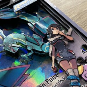 May include: A 3D pop-up card featuring a blue and green Pokemon character with a female character standing next to it. The card has a holographic background and the text 'on Pulse' and 'our deck' is visible. The card also features a pink Pokemon character in the background.