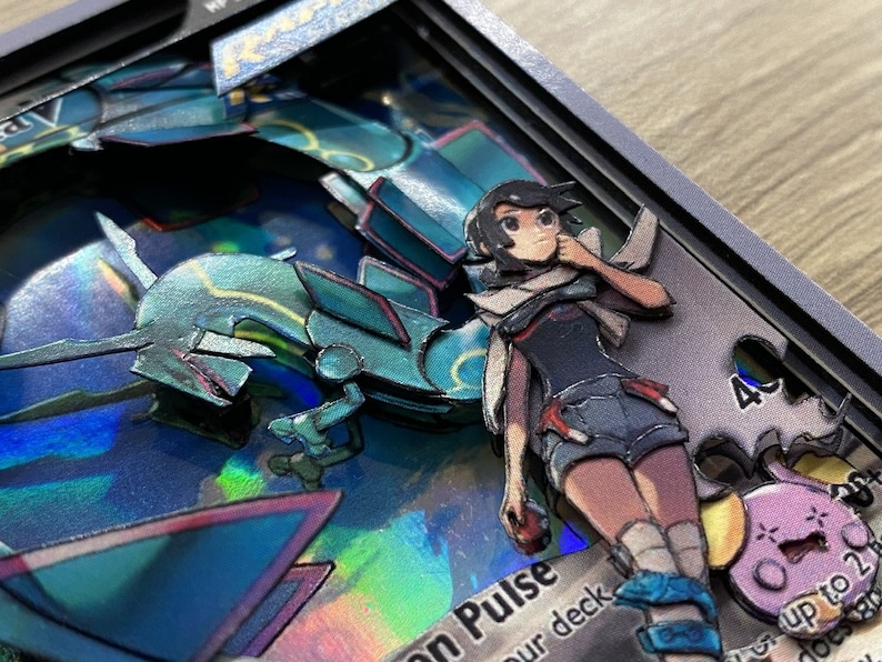 May include: A 3D pop-up card featuring a blue and green Pokemon character with a female character standing next to it. The card has a holographic background and the text 'on Pulse' and 'our deck' is visible. The card also features a pink Pokemon character in the background.