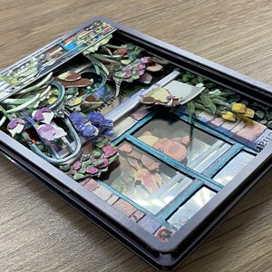 May include: A 3D pop-up card with a gray frame and a colorful scene inside. The scene features a window with a blue frame, flowers, and a small building with a sign that reads 'PlantDoc® Patent Pending'. The card also has a small label that reads 'BASIC'. 