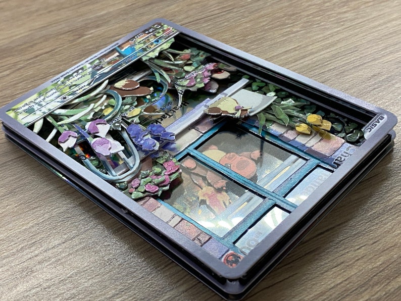 May include: A 3D pop-up card with a gray frame and a colorful scene inside. The scene features a window with a blue frame, flowers, and a small building with a sign that reads 'PlantDoc® Patent Pending'. The card also has a small label that reads 'BASIC'. 
