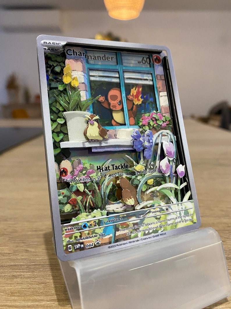 May include: A Pokemon trading card featuring Charmander, a fire-type Pokemon. The card is a 3D pop-up style with a detailed background of a window with flowers and plants. The card has a light blue background with the text 'BASIC' in the top left corner. The card has the text 'Charmander' in the top center. The card has the text '60' in the top right corner. The card has the text 'Hat Tackle' in the center. The card has the text 'This Pokemon can't be damaged by attacks that make it go to sleep.' in the center. The card has the text 'Weakness: Water x2' in the bottom left corner. The card has the text 'Resistance: None' in the bottom center. The card has the text 'Retreat: 1' in the bottom right corner. The card has the text 'From the moment it is born, a flame burns at the tip of its tail. It is said that the flame will go out if the Charmander's life force is ever to go out.' in the bottom center. The card has the text 'Illus. MINAMINAMI Toke' in the bottom left corner. The card has the text '©2023 Pokemon/Nintendo/Creatures/GAME FREAK' in the bottom right corner. The card has the text '6 SVP 044' in the bottom left corner.