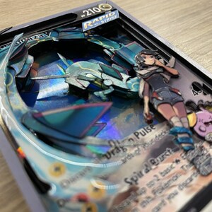 May include: A Pokemon card with a blue and silver holographic design. The card features a character with pink hair and a blue and white outfit. The card text reads 'Drag Pulse' and 'Spiral Burst'. The card is framed in a black and silver frame. The card is part of the Rapid Strike series.
