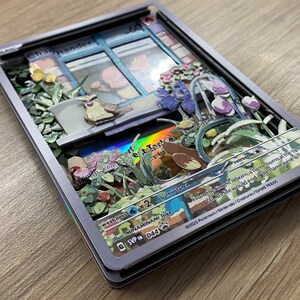 May include: A Charmander Pokemon card with a 3D pop-up design. The card features a window with a Charmander inside, surrounded by flowers and greenery. The card is silver with a holographic effect. The text on the card reads 'Charmander', 'Pokemon', 'Weakness 2', 'Resistance', 'Retreat', 'From the time it's born, a flame burns at the tip of its tail. Its little tail would end the flame were it to go out.' and '©2023 Pokemon/Nintendo/Creatures/GAME FREAK.'