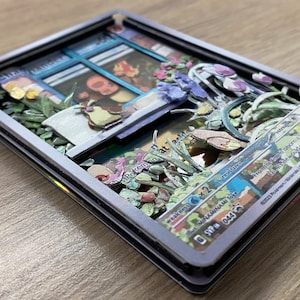May include: A 3D pop-up Pokemon card with a blue and white window frame, a green plant, and a brown bird. The card is surrounded by a silver frame and has the text 'Pokemon' and 'Weakness x2' on it. The card also has the text 'SVP 044' on it.