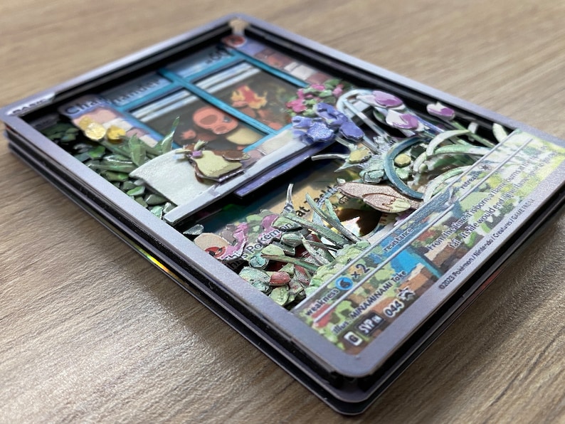 May include: A 3D pop-up Pokemon card with a blue and white window frame, a green plant, and a brown bird. The card is surrounded by a silver frame and has the text 'Pokemon' and 'Weakness x2' on it. The card also has the text 'SVP 044' on it.